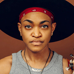 A head-and-shoulders portrait of Sug Daniels.  She is a young black woman with a strong, direct gaze.  She has thick arched eyebrows, a broad nose with a stud on one side, very full lips, and medium-brown skin. She is wearing a black hat with a broad brim, tilted up, so all we see is a wide black disc behind her head. The photo is cropped so the edge of the hat almost doesn’t show. She also has a red bandanna wrapped around her head, with an inch or so showing beneath the hat brim.