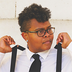 Crys Matthews, a young Black woman, wears a crisp white button-down men’s shirt, black tie and black suspenders. She has her thumbs underneath the suspender straps, pulling them up off her shoulders. Her curly hair is long on top and cropped close at the sides.