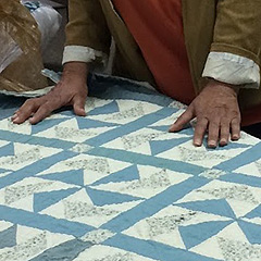 Narda LeCadre’s hands on a quilt. She is black, and her hands suggest she is an older woman. She is wearing a brown coat with its cuffs turned back, and a rust-colored top. The quilt has blue calico pinwheels.