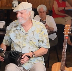An older man in a tropical shirt and peaked cap plays a concertina.