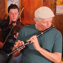 In the foreground, an older man wearing a cap plays a wooden flute. Behind him, a young woman is playing a fiddle.