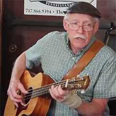 An older man wearing a cap plays guitar and sings a song at the session.
