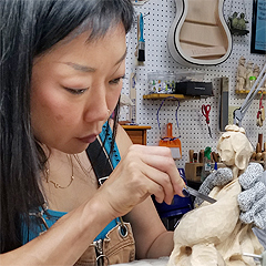 This closeup shot focuses on Aron Rook’s face and hands, and the wood sculpture she is carving. Aron is a young woman of Asian appearance, with straight black hair. She holds her piece in one gloved hand and removes material with a small tool.