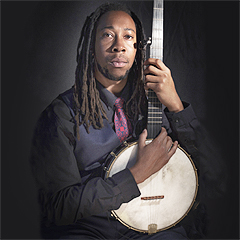 Hubby Jenkins holds a banjo in his lap with the fretboard vertical.  He is a Black man with dreadlocks, wearing black shirt, vest, and a patterned red tie.