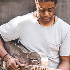 King Jester is a young Black man, leaning against a wall as he plays a metallic resonator guitar. He wears a gray T-shirt and is looking down at the guitar.