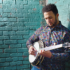 A Black man stands in the corner of a brick-walled room, playing a banjo and gazing down toward his hands.