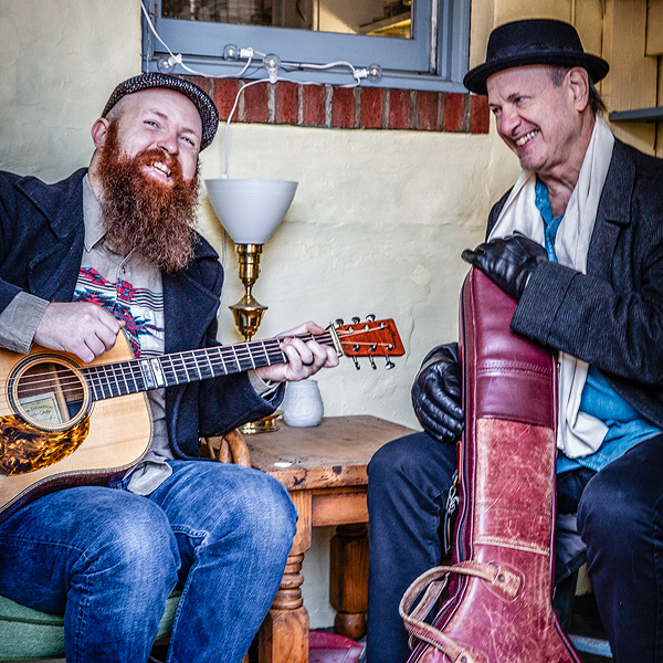 Bryan McDowell & Mark Schatz sit on a porch. McDowell holds a guitar; Schatz holds a banjo case.