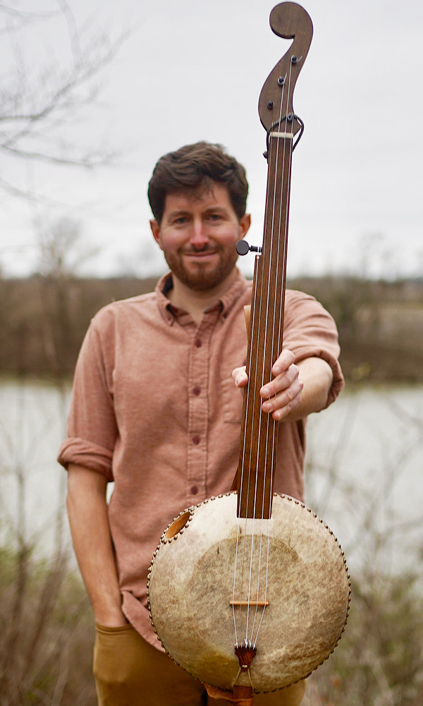 The gourd banjo, held up for inspection. Brad Koloder is a young man with dark hair and beard, standing outdoors.