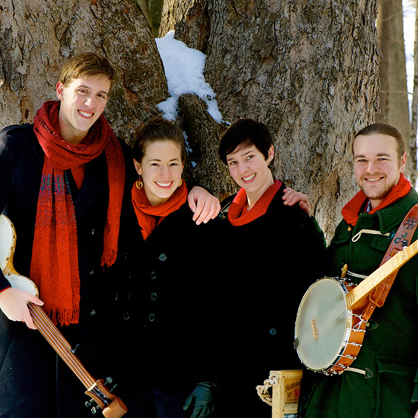 The four singers of Windborne, outdoors on a winter day. The two young men (holding banjos) and two young women are all wearing black coats and red scarves, in front of an enormous tree with snow in a cleft just above their heads.