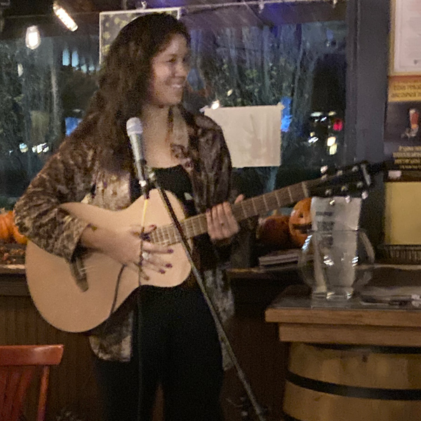 Eryn Spangler performs at a local bar. She is holding a guitar and grinning. 