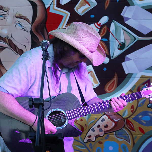 Joe Cooney performs in front of a colorful mural, playing a black cutaway guitar.