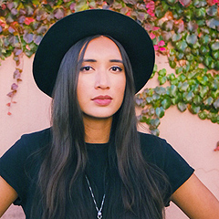 A head-and-shoulders shot of Raye Zaragoza, a young multi-racial woman with long straight hair, dusky skin and full lips.  She is wearing a black hat (only the underside of the brim is seen) and black T-shirt. Her expression is serious and a little sad.