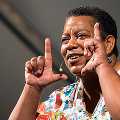 Sheila Arnold, an older Black woman with very short hair, holds her fingers up in L shapes, like a frame that she is looking through. She smiles as she speaks to her audience.