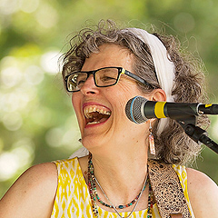 Jamie Anderson performing onstage, laughing and looking off to her right. She is playing a guitar and wearing a yellow-and-white dress with open shoulders.  Her curly graying hair is held back from her face with a wide white hair band.  She is wearing dark-framed glasses and four necklaces.