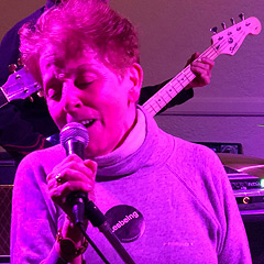 Kay Turner strikes a rocker pose, lit up with magenta light and singing into a microphone on a stand that she's pulled over at an angle. Behind her are people playing electric bass and rhythm guitar, but only the headstocks are visible. The rock & roll pose is at odds with her grandmotherly appearance with loosely curled short hair and turtleneck sweatshirt. She's wearing a large black button with white print that says 'Lesbing.'