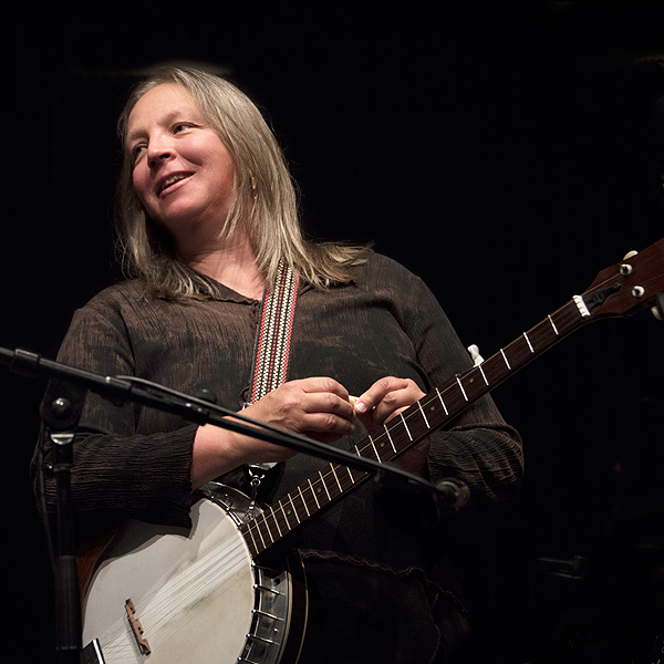 Barb Barton performing onstage, smiling and looking off to her right as if speaking to someone. She has a banjo on a colorful neckstrap but her hands are held together above it. She is wearing a dark brown shirt that blends in with the very dark background. Her gray-blonde hair is straight and the feathered ends fall on her shoulders.