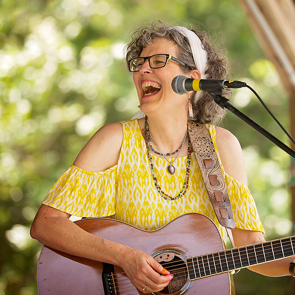 Jamie Anderson performing onstage, laughing and looking off to her right. She is playing a guitar and wearing a yellow-and-white dress with open shoulders.  Her curly gray-blonde hair is held back from her face with a wide white hair band.  She is wearing dark-framed glasses and four necklaces.