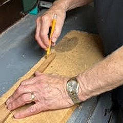 A close-up of a man's hands as he uses a pencil to trace around a template laid on a thick block of wood, to draw a cutting line onto the wood.