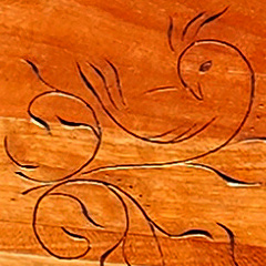 Closeup of one of Judy Walton’s carvings on a piece of cherry wood: a stylized bird outline among viney plant tendrils. The bird is looking over its shoulder and down, with its beak against the top of its wing.