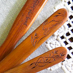 Closeup of three cherry-wood spatula handles against a handmade lace background.  Each is decorated with a simple line carving in the Pennsylvania Deutsch folk art style: tall wildflowers on one, a running rabbit on another, and words on the third.