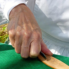 Closeup of Judy Walton’s right hand as she starts to carve a design in a spatula handle.  Her knife follows a pencil line draft on the wood. The wood is laid on a tabletop covered with a green cloth.