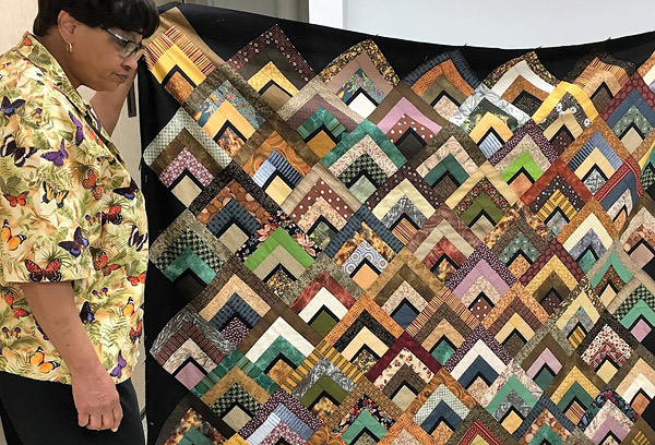 A Black woman, Narda LeCadre, shows off a quilt she made.  Its blocks are set next to one another with no framing, and are placed diagonally against a black background.  Made of strips in various earth colored prints, the blocks give the impression of overlapping hills or arrows.