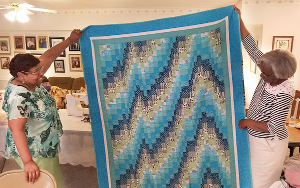 Marie Fourney (left) holds a corner of a quilt she made, with another Black Woman holding up an opposite corner. The design features bands of color zigzagging up from lower left to upper right.  The bands are shades of turquoise, with darker blue just below each band suggesting a shadow.  Darker grays and greens set off the bright bands. The quilt’s border is about 9 inches wide, blue at the outside framing narrower bands of white and sea-green.