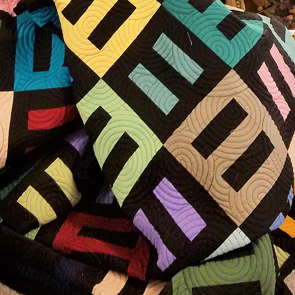 The quilt is piled loosely on a chair.  Its pattern is made up of blocks maybe 9 inches square, each one a large, bold ‘equals’ sign. The blocks alternate, turned at 90 degrees to one another: two vivid solid-color bars on a black ground, or two black bars on a colored ground. The concentric circles of quilting look like stacked discs.