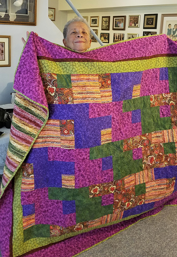 Cynthia’s quilt is mae up of four-inch squares, sewn together in irregular color groups -- violet, royal blue, green, and various brown shades. The quilt obscures all of Cynthia but her proudly smiling face. She is African American with short grey hair and a broad face.