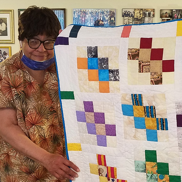 Narda LeCadre holds up a quilt she made. The quilt is mostly white, with diagonal lines of small blocks of colorful fabrics.  Narda is a Black woman with short hair and dark-rimmed glasses, and she is smiling proudly.