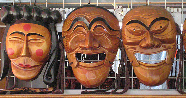 Three Talchum masks hang on a wire grid. The mask at left appears to be a woman, with shoulder-length hair and a horizontal roll of twined hair across the top. Quarter-sized red dots are painted on cheeks and forehead, and the center of the lips are painted the same red. Her eyes are closed and she seems to be smiling.  The center mask is of a laughing man with prominent eyebrows and eyes squinched shut.  The mask at right is of a bald man with wide-open eyes and wide-open semi-smiling mouth.
