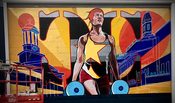 A mural on the side of a building, depicting a woman athlete with short red hair. She is holding dumbells in each hand, wearing red shorts and a gray tank shirt with a large gold graphic star covering half her chest.  Line drawings of urban buildings are to her left and right.  Behind her, there are rays of gold sunlight, and a large orange disc over her shoulder on the right side.