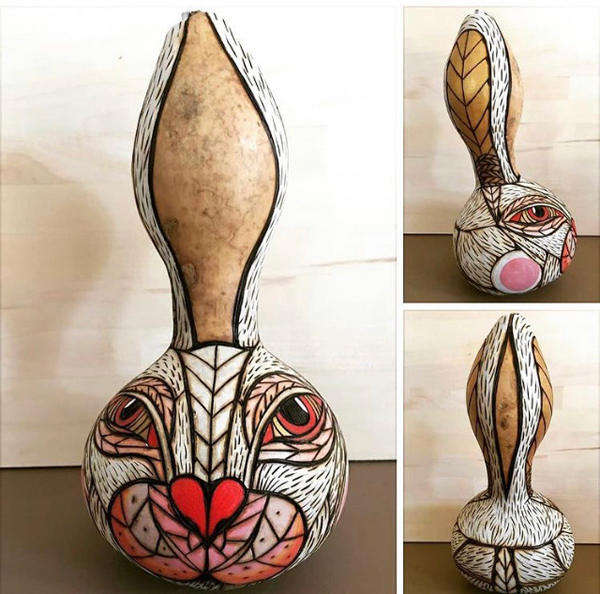 A gourd that has been decorated (with wood-burning) as a stylized rabbit.  The round part of the gourd is its face -- with a heart-shaped nose, rather human eyes, and hash-marks indicating fur. The rabbit’s ears go up the sides of the gourd’s tall neck