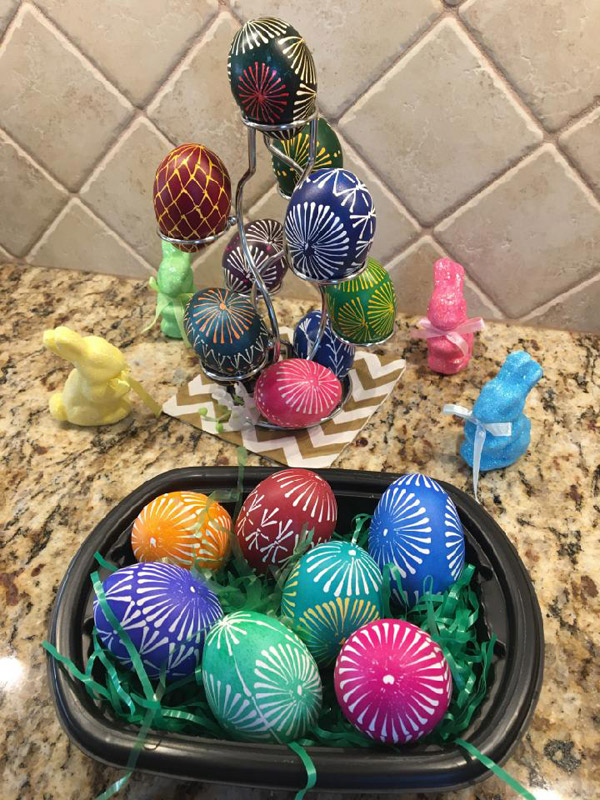 In the foreground: a black plastic bowl with a little plastic ‘Easter grass’ holds seven decorated eggs in red, green, blue, purple, orange and maroon.  Most of the eggs feature variations on a ‘pinwheel’ pattern that resembles the spokes of a bicycle wheel radiating from a central dot.  Most of the eggs have four of these pinwheels circling the egg, with smaller pinwheels on top and bottom. Behind the bowl, a wire ‘egg tree’ holds more decorated eggs in deeper colors, with some multicolored designs. Small bunny statues complete the display.