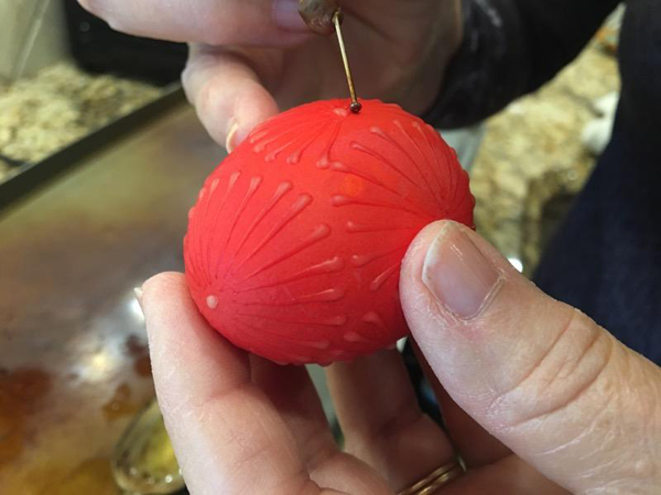 Closeup of Barbara drawing a traditional pinwheel design on an egg that’s dyed red. She’s using her pin-on-a-pencil tool to apply melted wax in the “drop and pull” method