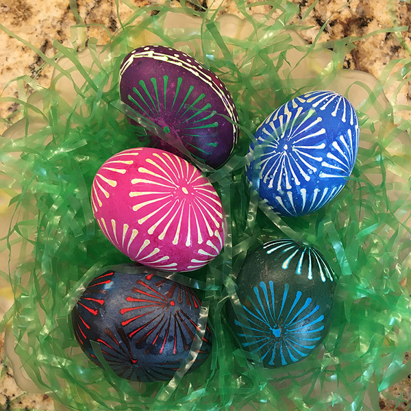 Five decorated eggs in pink, blue, green, red and maroon rest on a pile of plastic ‘Easter grass’.  Most of the eggs feature variations on a ‘pinwheel’ pattern that resembles the spokes of a bicycle wheel radiating from a central dot.