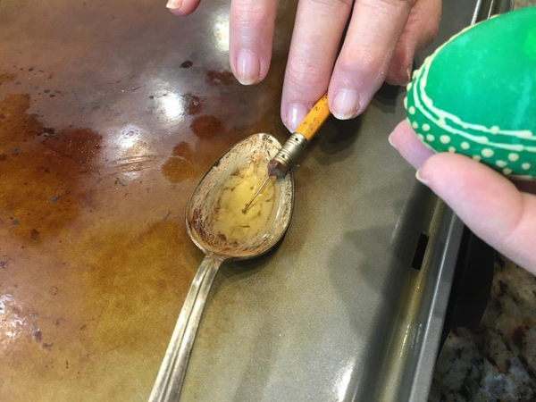 In the center of this shot, we see an large silver spoon with a rather battered and scratched bowl.  It’s on a flat heated griddle, and contains a small amount of melted wax.  Coming in from the top of the image, we see Barbara’s fingers holding a pencil lightly. A pin is stuck into the pencil’s eraser, and the head end of the pin is dipped into the melted wax.  On the right edge of the image, we see a green egg that Barbara is going to apply the wax to.
