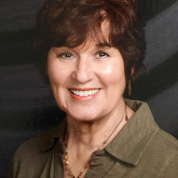 Barbara Felty smiles for a portrait.  She has an oval face, bright brown eyes and wavy short brown hair. She appears to be in her 50s or 60s. She is wearing an open collared shirt of olive heather fabric, and a necklace of various-size amber beads on a bronze chain.