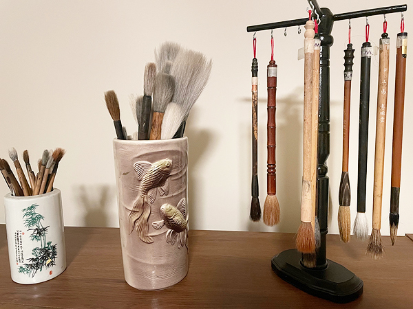 Diana’s stock of brushes.  At right, eight brushes hang hair-end-down from the crosspieces of a wooden stand.  To the left, a small cup holds smaller brushes and a larger cup holds larger brushes standing hair-end-up.