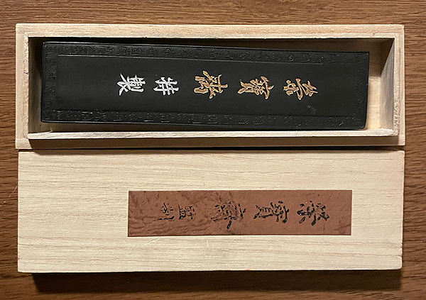 A narrow wooden box holds an ink stick, which appears to be about six inches long by one inch wide, and maybe half an inch thick.  The ink stick is black and is embossed with a frame and five gilt Chinese characters.