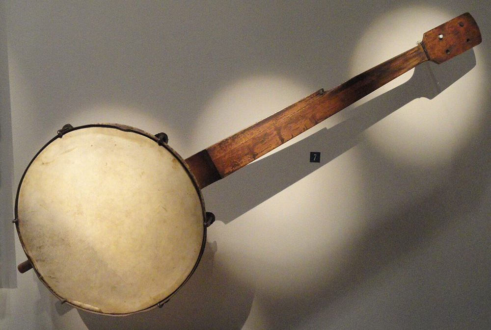 A battered antique banjo. The skin head is intact, but the ring is dented in several places, and the tuning pegs are missing. Its fretboard is arranged for four long strings and one short string, but the strings are not present. The tensioning ring has six hooks.