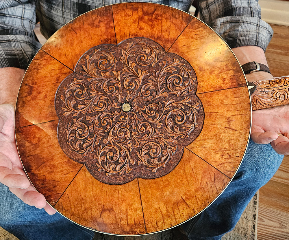 Glenn holds a closed-back banjo he made.  The back has a medallion of intricately carved dark wood in the center. Surrounding that are eight wedges of a lighter stained wood with interesting graining. The heel piece (where the fretboard joins the round part of the banjo) is also intricately carved.