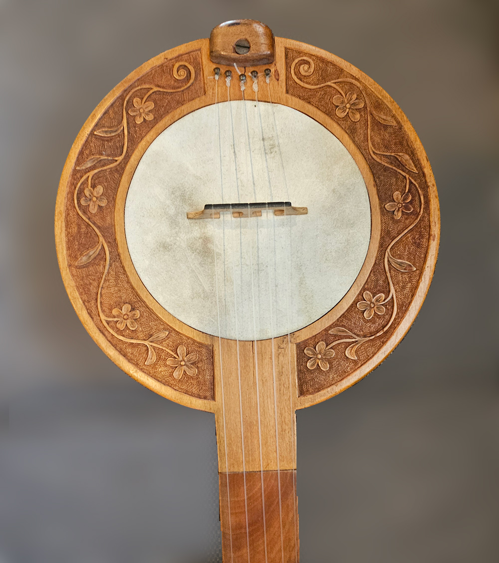 The round part of this banjo is two concentric discs: a skin head in the center, perhaps 8 inches in diameter, and around that is an engraved wooden ring, perhaps 1.5 inches wide. The ring is engraved with a flowering vine against a darker textured background, simple but not primitive. Five strings are stretched across, with a bridge at the center of the skin head area. The strings continue up a fretless fretboard, but the photo does not show where they end.