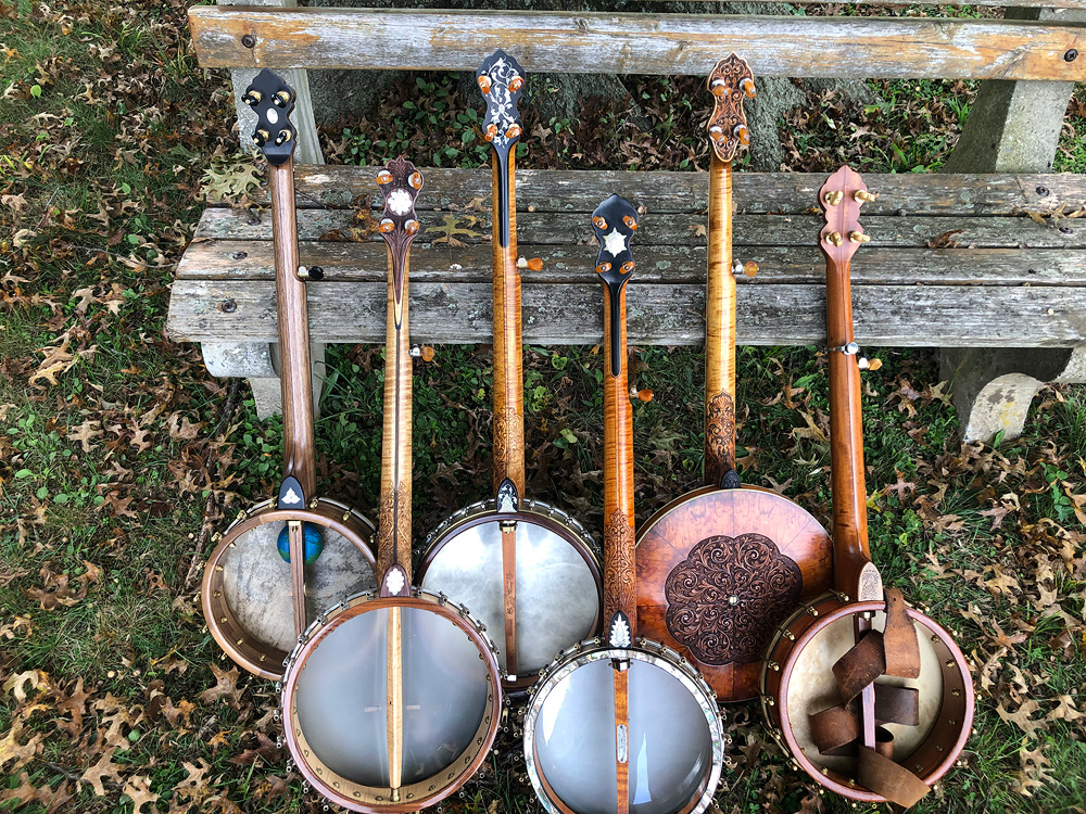 Six banjos made by Glenn, leaned against a weathered wooden bench to show decorations visible from the back. One has a carved and inlaid resonator; the others are open-back.