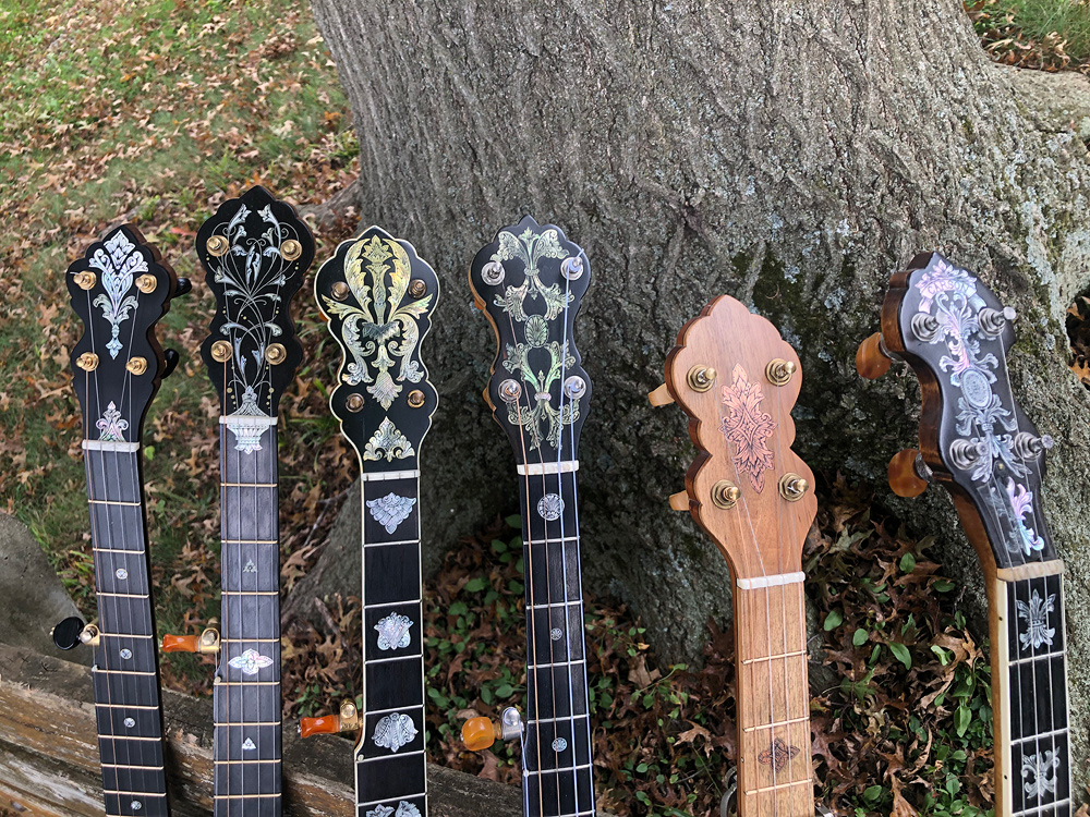 Elaborately-inlaid pegboards and inlaid fretboards of six banjos made by Glenn, leaned against a weathered wood fence with a large tree behind it.