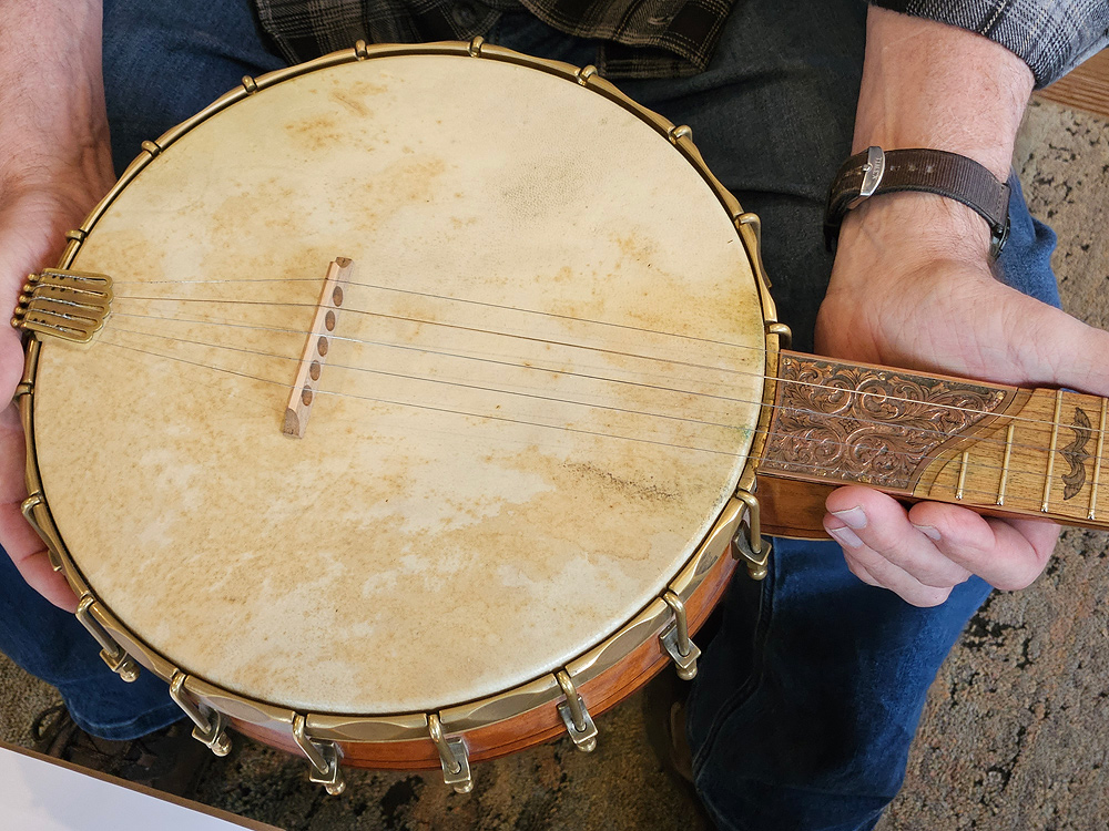 Glenn holds a banjo he made, showing the hooks, scallops and scoop described above. A sturdy brass ring encircles the banjo head, holding the skin head onto the wooden frame ring. Around the edge, every two inches or so, a hook-and-screw mechanism attaches the brass ring at the top to the bottom edge of the wood frame. Between each of those hooks, the brass ring has some material beveled away, creating a scalloped effect. The “scoop” is a flat plate on the fretboard where it joins the head, intricately carved and with a graceful arch at the end nearer the pegs.