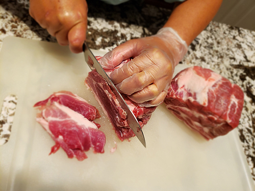 Ho-Thanh slices fist-size hunks of pork shoulder into thin slices on a cutting board. She is wearing a glove on the hand holding the hunk she’s slicing, and using a long, wide knife.
