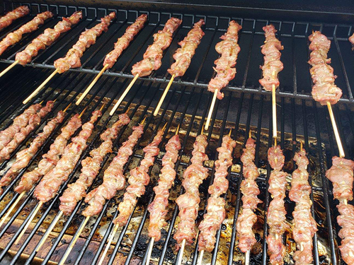 Skewers laid side by side to cook on a grill. There are 15 skewers on the main grill, and at least ten more on the smaller, upper shelf.