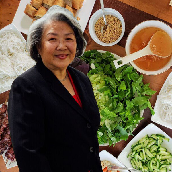 An image of Ho-Thanh Nguyen is superimposed over a display of Vietnamese foods ready to be assembled into Ho-Thanh’s signature dinner of Grilled Pork.  Ho-Thanh is a Vietnamese woman who appears to be in her 60s.  She has short graying hair that waves stylishly back from a broad oval face. She is smiling serenely with her eyebrows a little lifted.  She is wearing a black blazer over a barely-visible v-necked red blouse.