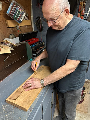 Stewart Walton stands at his workbench, preparing a slab of cherry wood to be cut into spatulas. He is drawing around a template with a pencil, marking the outline onto the wood. He appears to be in his 70s, and is looking down at his work.